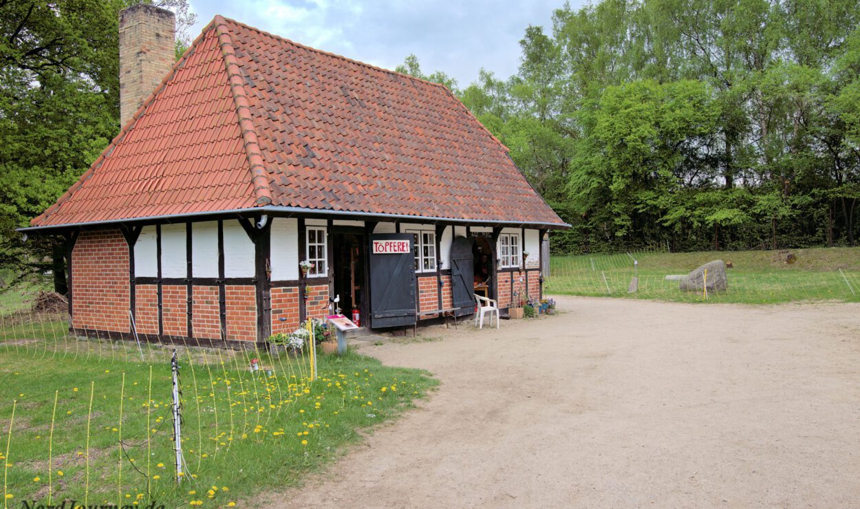 Ein kleines, traditionelles Haus aus Backstein und Holz mit rotem Ziegeldach steht in einer ländlichen Gegend, umgeben von Gras und Bäumen. Über der Tür hängt ein Schild mit der Aufschrift „KØBTE“.
