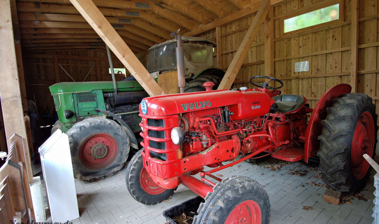 Zwei Oldtimer-Traktoren, einer rot und einer grün, sind in einer Holzscheune mit Fliesenboden und Holzbalken geparkt.