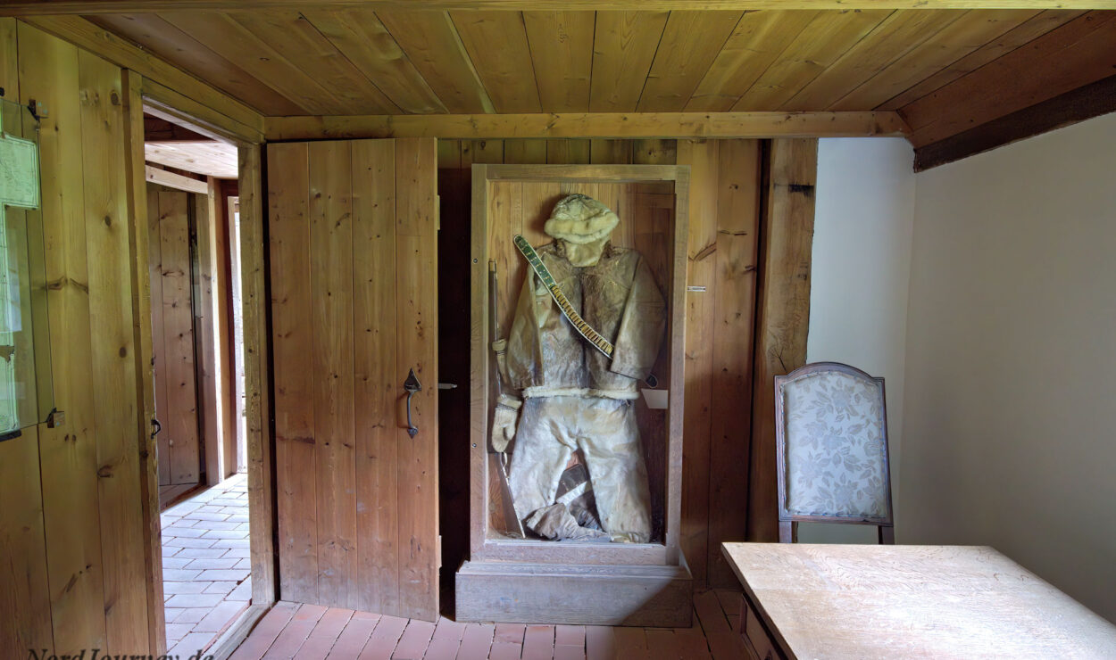 Eine Holzstatue eines Bergmanns steht in einer Holznische in einem rustikalen Raum mit Holzwänden, einem einzelnen Stuhl und einem kleinen Tisch.