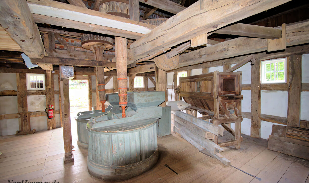 Innenansicht einer alten Holzmühle mit großen Zahnrädern, freiliegenden Balken, Mahlgeräten und natürlichem Licht, das durch kleine Fenster einfällt.