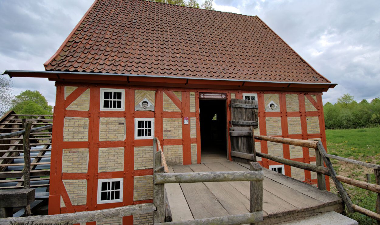 Eine traditionelle Fachwerk-Wassermühle mit Holzrampe, rotem Ziegeldach und offener Tür, eingebettet in eine grüne ländliche Landschaft unter einem bewölkten Himmel.