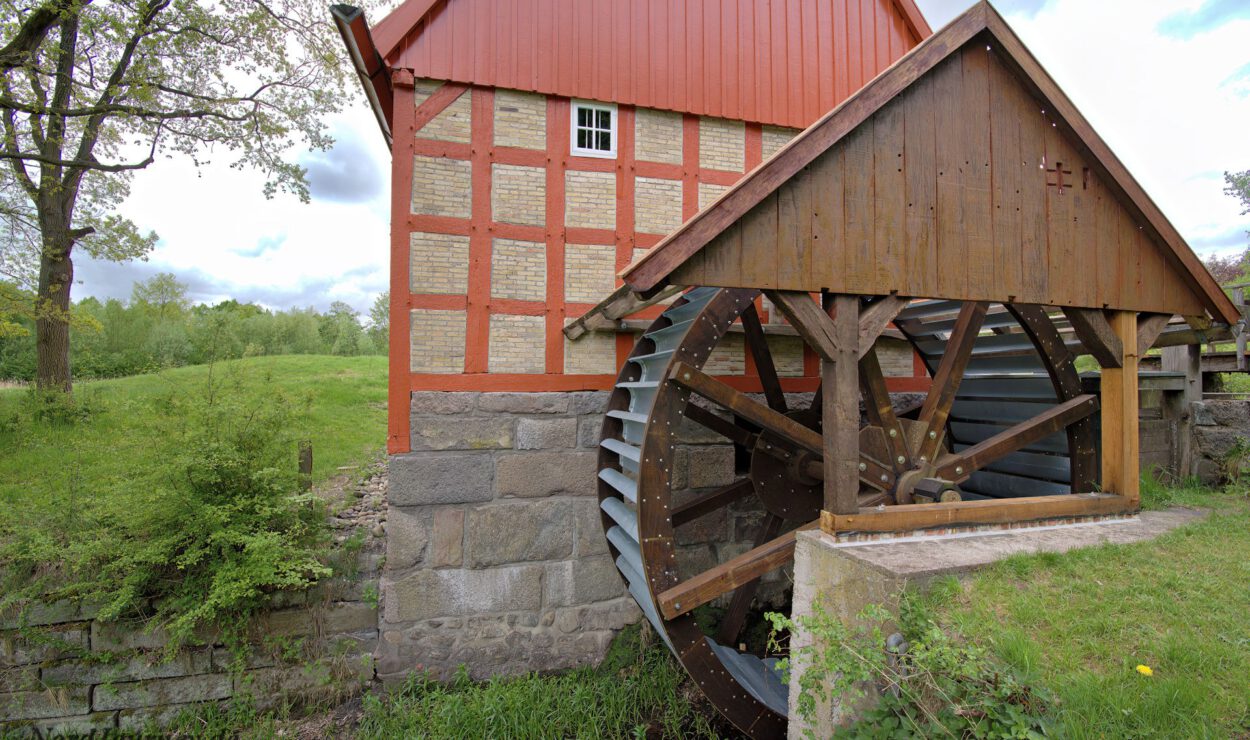An der Seite eines traditionellen Gebäudes aus Ziegeln und Holz ist ein großes hölzernes Wasserrad angebracht, umgeben von Gras und Bäumen.