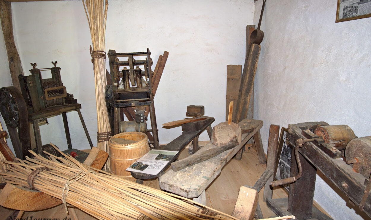 In einem Raum werden alte Holzbearbeitungswerkzeuge, Bänke, Schilfbündel und alte Maschinen ausgestellt, die an eine historische Werkstatt oder einen Handwerksbereich erinnern.