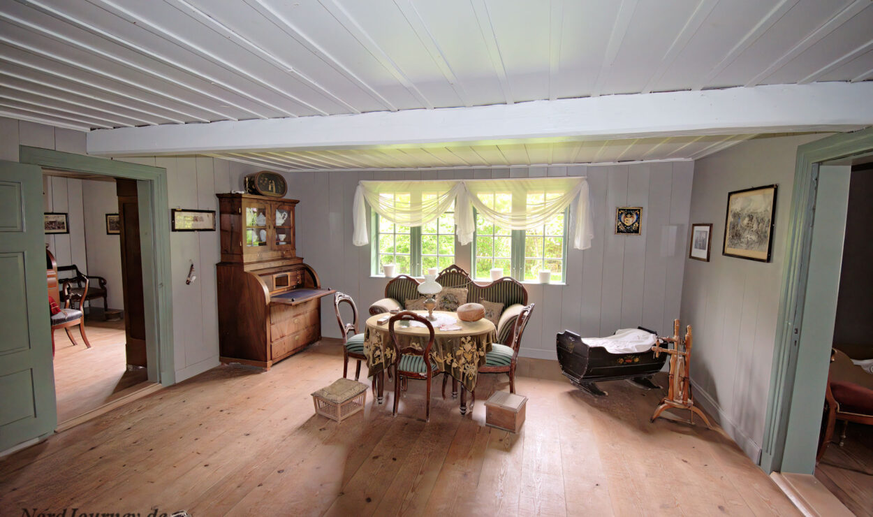 Ein Wohnzimmer im Vintage-Stil mit Holzböden, antiken Möbeln, einer Schaukelwiege, einem Spinnrad und Sonnenlicht, das durch die Fenster mit Vorhängen strömt.