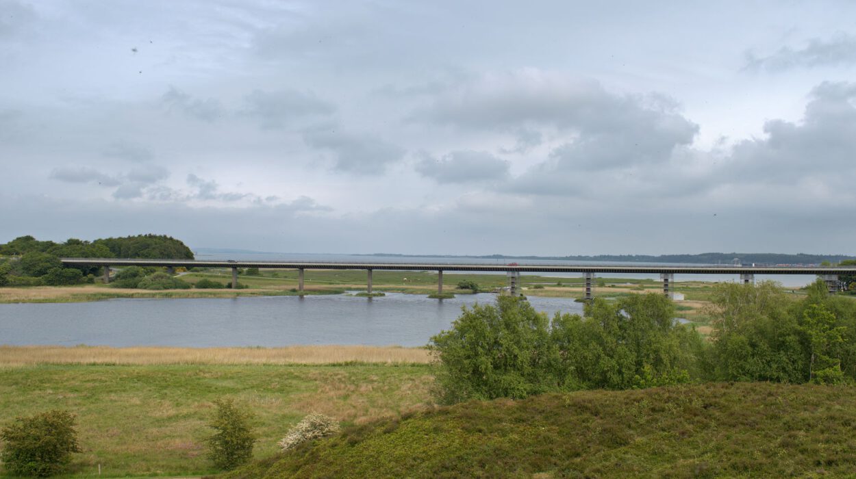 Eine lange Brücke überspannt einen breiten Fluss, umgeben von Wiesen und Bäumen unter einem bewölkten Himmel.