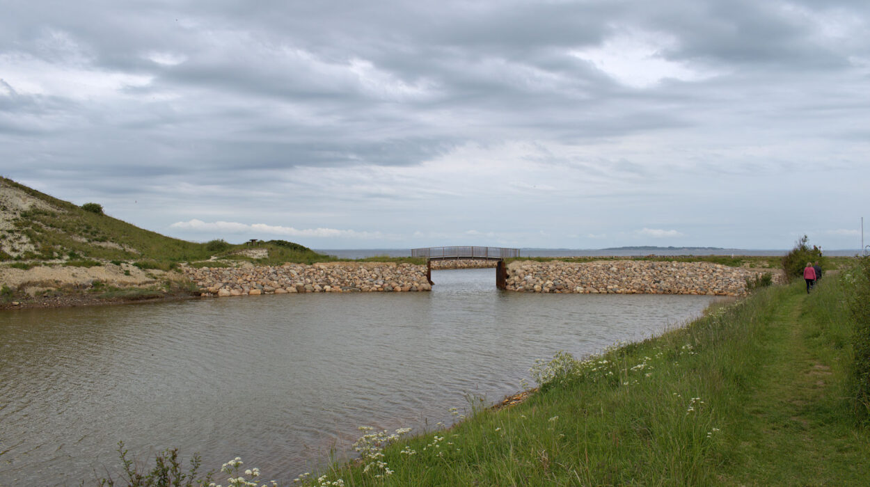 Eine kleine Steinbrücke überquert einen schmalen Fluss unter bewölktem Himmel, mit grasbewachsenen Ufern und einer Person, die auf einem Fußweg rechts davon geht.