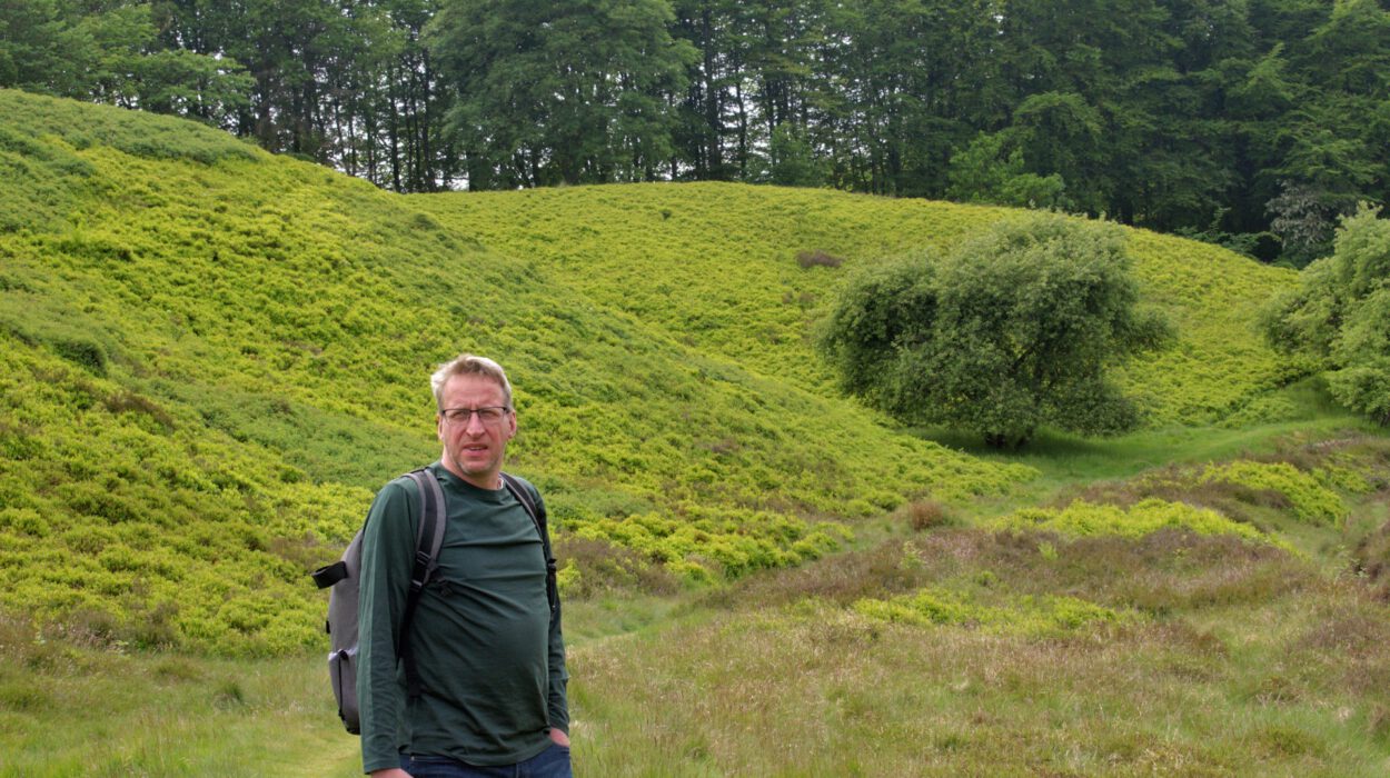 Ein Mann mit Brille und Rucksack steht in einer grasbewachsenen, hügeligen Landschaft mit Bäumen und dichter grüner Vegetation im Hintergrund.