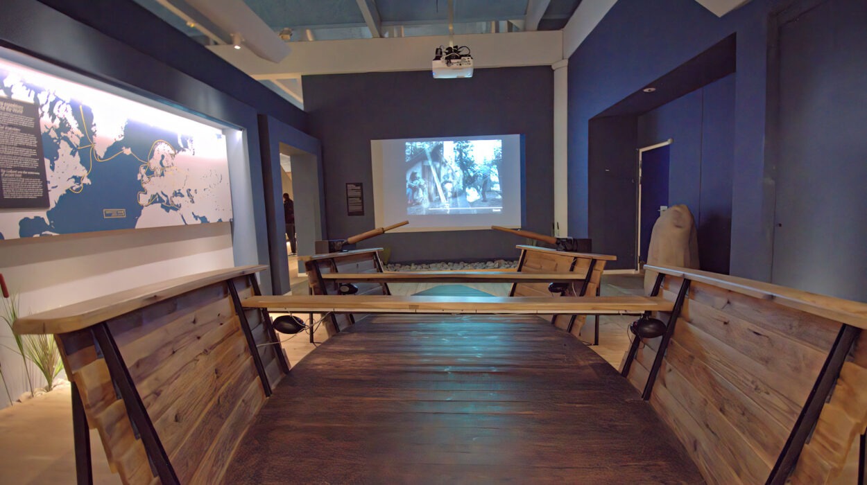 Eine Museumsausstellung mit Holzbänken in Form eines Bootes, die in einem modernen Raum mit blauen Wänden vor einem projizierten historischen Video auf einer Leinwand stehen.