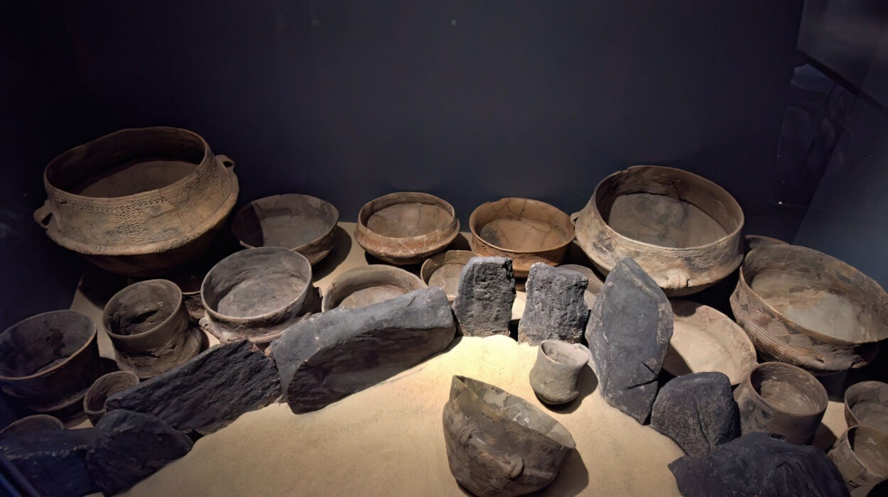 Eine Sammlung antiker Tongefäße und Schalen, die auf Sand angeordnet und von großen dunklen Steinen umgeben sind und in einem Museum ausgestellt werden.