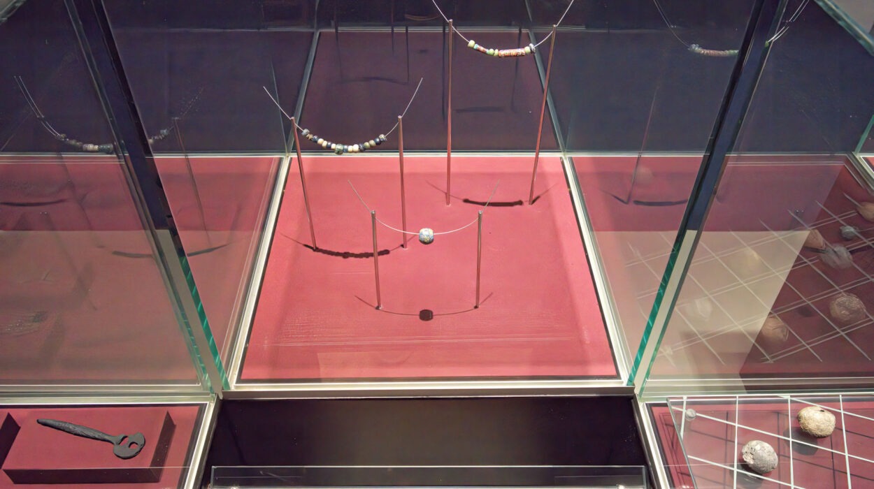 In einer Museumsvitrine sind antike Perlen und eine an Drähten aufgehängte Murmel ausgestellt, mit Informationstafeln, die die Gegenstände und ihre historische Verwendung beschreiben.