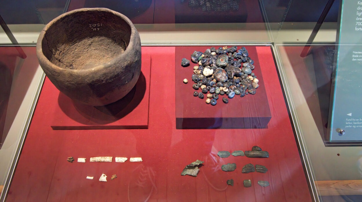 Ein großer antiker Tontopf, eine Sammlung von Perlen und kleinen Artefakten sowie mehrere Stein- und Knochenfragmente, die in einer gläsernen Museumsvitrine auf einer roten Fläche ausgestellt sind.